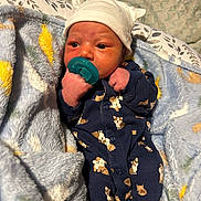 Amari is registered to the contest to win money with this photo: newborn, baby, pacifier, onesie, hat, blanket, infant, cute, sleepwear, cozy, soft, patterned_clothing, clothing, face, person, hand, bed, resting, indoor, child