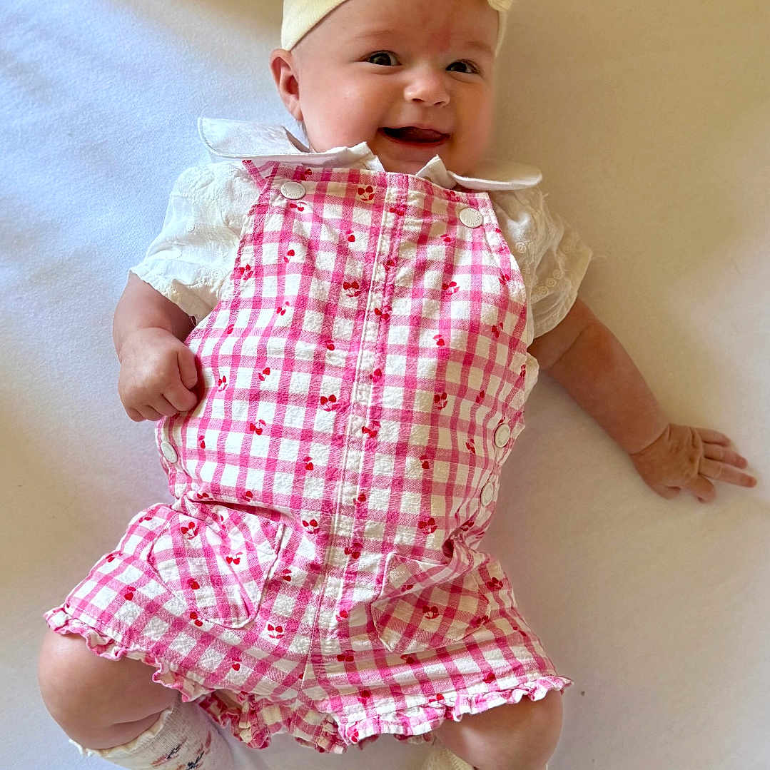 Constance participe au concours pour gagner de l'argent avec cette photo : baby, baby_clothes, cheerful, child, crib, cute, happy, headband, headwear, indoors, infant, lying_down, person, pink_clothing, romper, small_hands, smiling, socks, soft_lighting, white_sheet