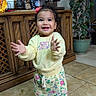 child, toddler, smiling, happy, standing, clapping, portrait, indoors, home, kitchen, wooden_cabinet, coffee_machine, houseplant, floral_clothing, sweatshirt, sneakers, pink_hair_bow, tile_floor, full_body, natural_light