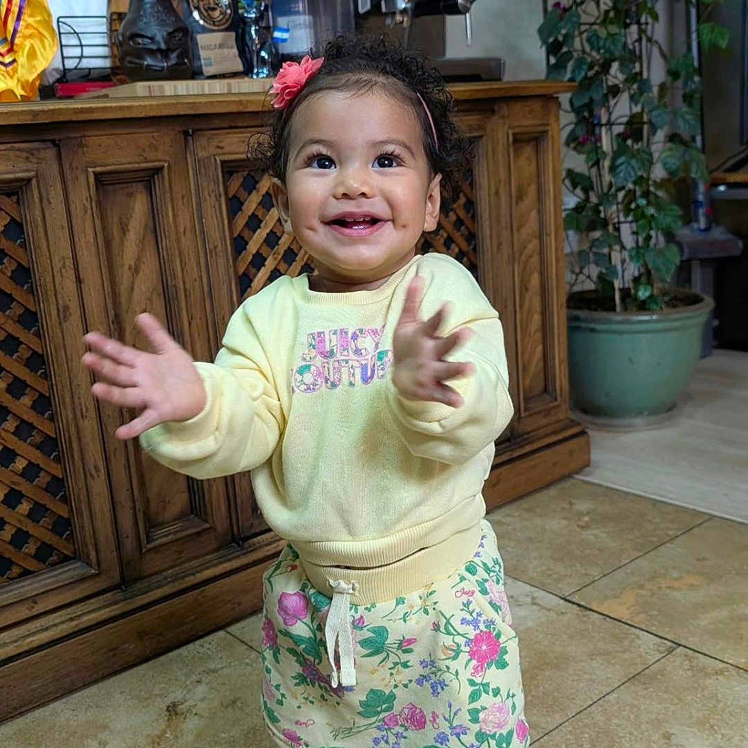 Lianna is registered to the contest to win money with this photo: child, toddler, smiling, happy, standing, clapping, portrait, indoors, home, kitchen, wooden_cabinet, coffee_machine, houseplant, floral_clothing, sweatshirt, sneakers, pink_hair_bow, tile_floor, full_body, natural_light