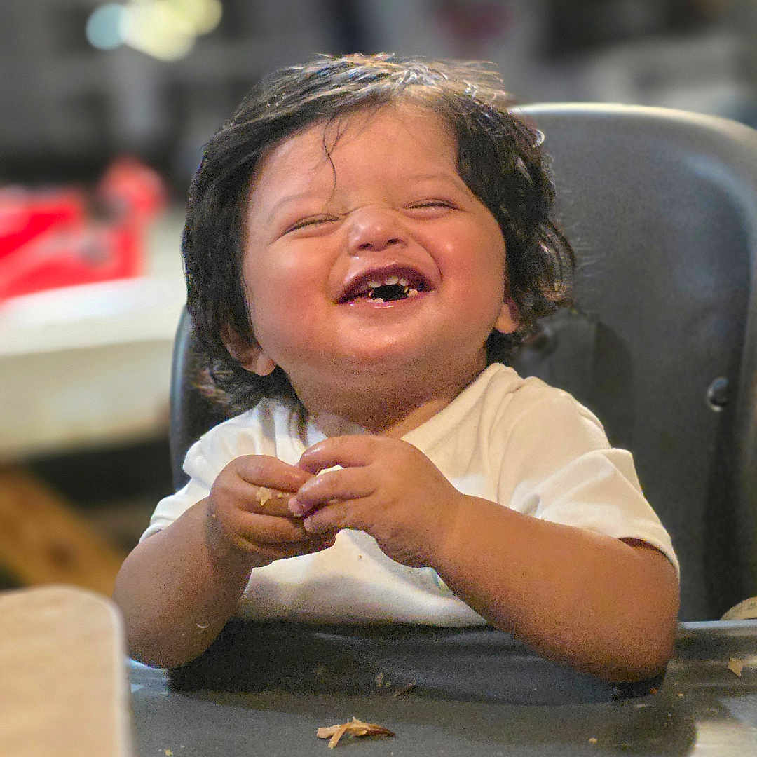 Moses joined the competition — help win amazing prizes! toddler, child, high_chair, food, messy, happy, laughing, indoor, meal_time, cute, smiling, baby, face, hands, table, hair, white_shirt, expression, person, seated