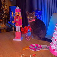 Bean is registered to the contest to win money with this photo: cat, nutcracker, table, christmas_tree, decorations, pink, blue_light, holiday, indoor, furniture, toy, heart, collar, chair, ornament, festive, glowing_eyes, wood, sitting, text_overlay