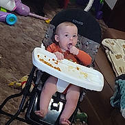 Koda is registered to the contest to win money with this photo: baby, child, infant, walker, highchair, snack, eating, hand, face, portrait, sitting, indoor, living_room, carpet, couch, toys, playroom, orange_shirt, barefoot, cosco