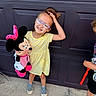 child, girl, glasses, yellow_dress, plush_toy, minnie_mouse, smiling, standing, outdoor, concrete, garage_door, playful, happy, sneakers, hair_up, person, toy, casual, summer, friends