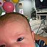 baby, close_up, face, striped_clothing, balloons, indoor, living_room, furniture, plant, television, high_chair, colorful, child, portrait, smiling, happy, cute, person, infant, home