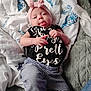 baby, infant, headband, bow, tongue, black_shirt, striped_pants, bed, blanket, white, blue, pattern, lying_down, child, cute, playful, comfortable, soft, indoors, person