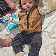 Xavier is registered to the contest to win money with this photo: baby, infant, hoodie, vest, sneakers, blanket, cat, orange_cat, person, arm, tattoo, hand, clothing, indoor, bed, soft_texture, cute, casual, resting, home
