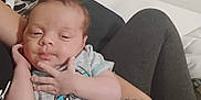 Xavier is registered to the contest to win money with this photo: baby, infant, onesie, sleepy, hand, face, person, adult, arm, clothing, bed, blanket, legs, tattoo, skin, resting, indoors, casual, holding, comfort