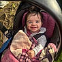 baby, infant, child, stroller, car_seat, blanket, smile, happy, blue_eyes, dark_hair, hand, hoodie, seatbelt, outdoors, grass, sunlight, portrait, patterned_blanket, tree_roots, cozy