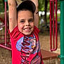 child, boy, smiling, playground, red_shirt, baseball_cap, monkey_bars, park, outdoor, trees, wood_chips, face, teeth, arms_up, casual_clothing, play, batman, happy, portrait, depth_of_field