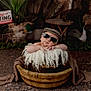 Colton is registered to the contest to win money with this photo: baby, sunglasses, cap, wooden_bowl, fluffy_fabric, rustic, sign, gone_hunting, greenery, outdoor, decor, relaxed, cute, child, portrait, hat, brown_blanket, floor, person, fashion