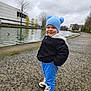 toddler, child, blue_hat, black_jacket, blue_pants, sneakers, outdoor, canal, pathway, overcast, trees, water, urban, smiling, cute, winter_clothing, standing, person, playful, park