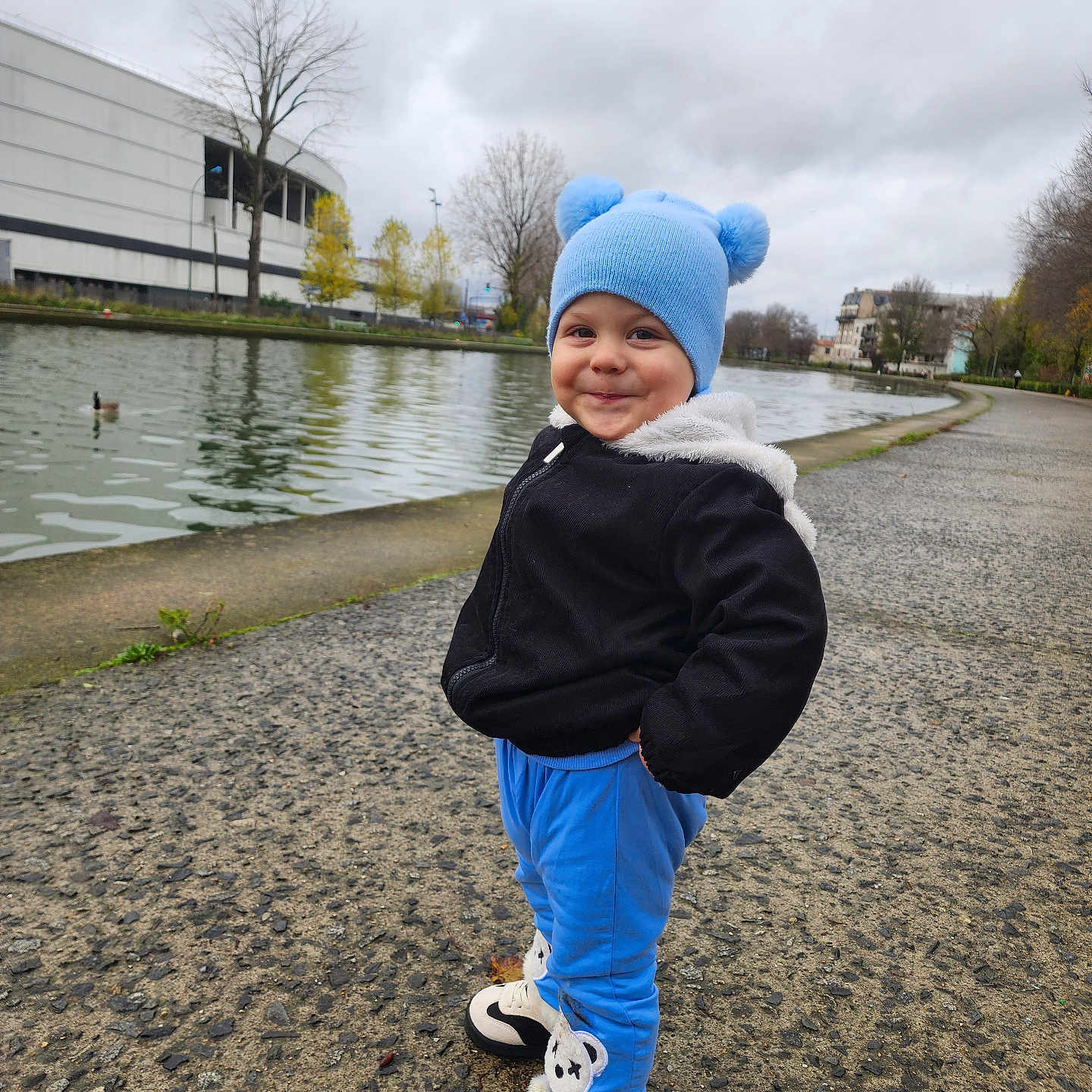 Mathias-Gabriel participe au concours pour gagner de l'argent avec cette photo : black_jacket, blue_hat, blue_pants, canal, child, cute, outdoor, overcast, park, pathway, person, playful, smiling, sneakers, standing, toddler, trees, urban, water, winter_clothing