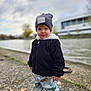 toddler, child, outdoor, river, cloudy_sky, hat, jacket, pants, sneakers, sidewalk, building, nature, autumn, leaves, casual_clothing, portrait, walking, young_child, urban, daytime