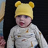 baby, child, yellow_hat, bear_ears, animal_print, shirt, pants, blue_eyes, smiling, face, hand, chair, cushion, indoor, curious, seated, cute, playful, infant, clothing