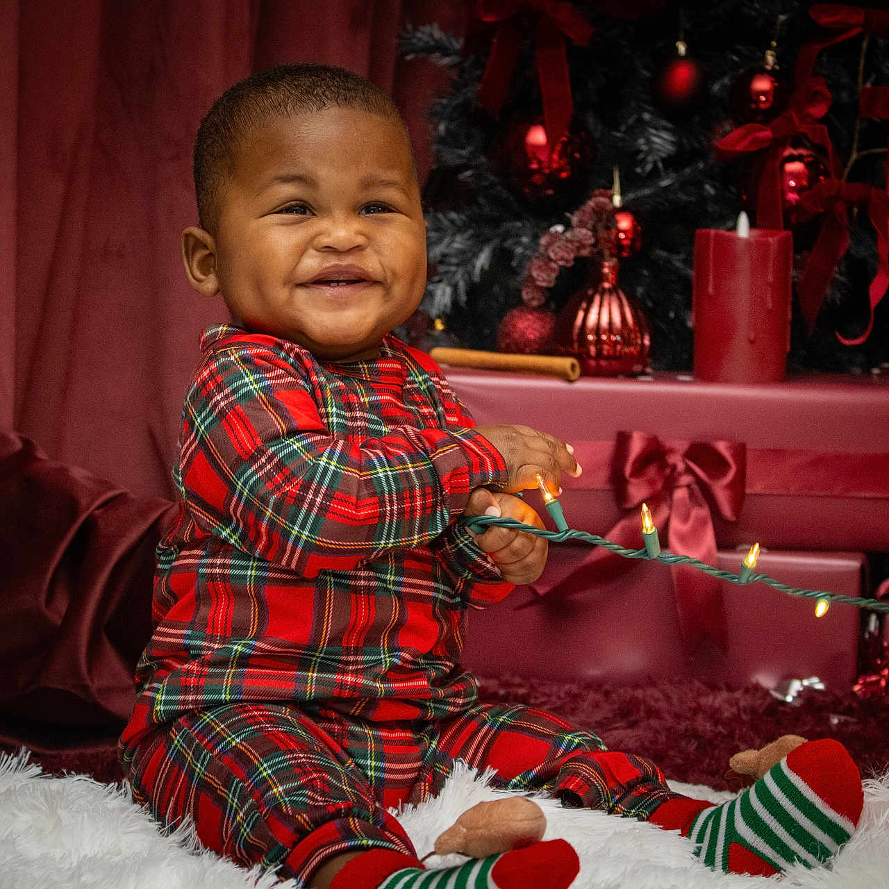 Kingston is registered to the contest to win money with this photo: baby, child, smiling, plaid_pajamas, christmas_lights, holiday, presents, red, decorations, candle, christmas_tree, ribbon, festive, indoor, celebration, happy, cute, sitting, cozy, toy