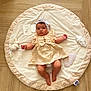 baby, infant, child, dress, headband, plush_toy, milestone_card, play_mat, wooden_floor, soft_toy, cute, lying_down, indoors, portrait, clothing, baby_girl, smiling, decor, fabric, person