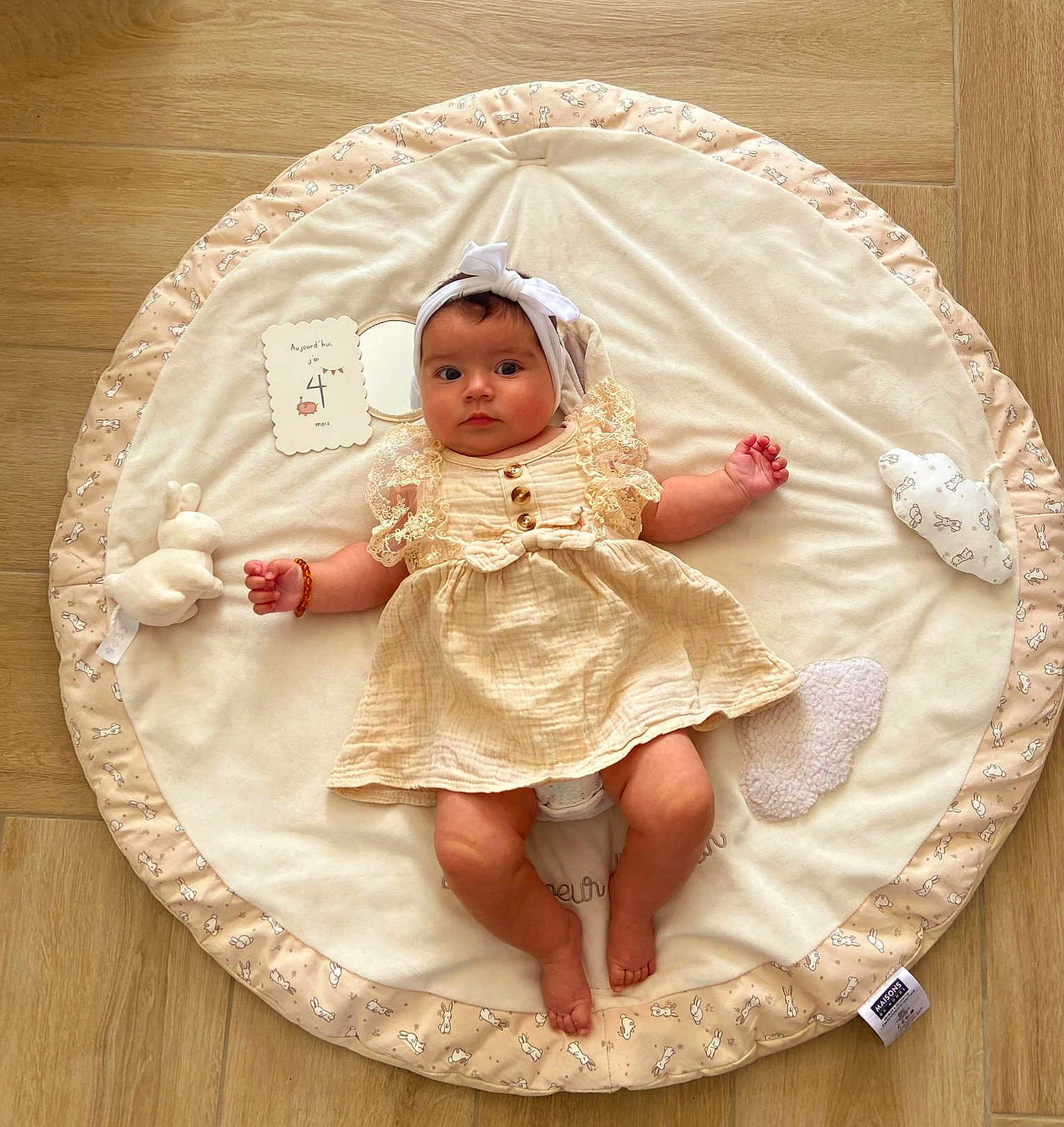 Izïa a rejoint le concours — aidez-le/la à gagner de superbes lots ! baby, infant, child, dress, headband, plush_toy, milestone_card, play_mat, wooden_floor, soft_toy, cute, lying_down, indoors, portrait, clothing, baby_girl, smiling, decor, fabric, person