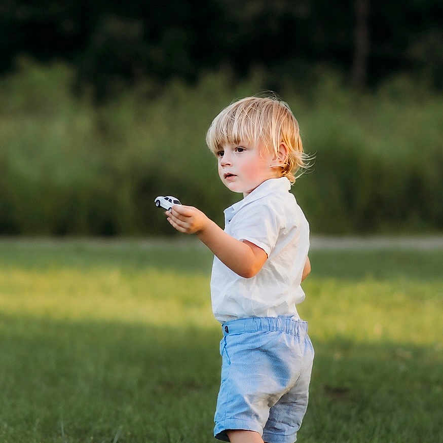 Otto is registered to the contest to win money with this photo: child, toddler, blonde_hair, toy_car, grass, outdoor, barefoot, shorts, white_shirt, summer, nature, greenery, playing, young_child, standing, side_view, daylight, casual_clothing, portrait, serious_expression