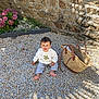 toddler, child, smiling, outdoor, stone_wall, stones, bag, woven_bag, flowers, sunlight, shadow, pants, sweater, barefoot, nature, garden, happy, sitting, playful, daytime