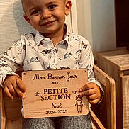 Naël participe au concours pour gagner de l'argent avec cette photo : child, boy, smiling, shirt, wooden_sign, chair, indoor, portrait, face, hands, clothing, wood, furniture, happy, cute, person, casual, home, background, young