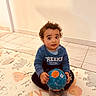 ball, blue_sweater, casual_clothing, child, curious, curly_hair, cute, floor_mat, home, indoor, looking_up, person, play, play_mat, sitting, smiling, tile_floor, toddler, toy, young_child