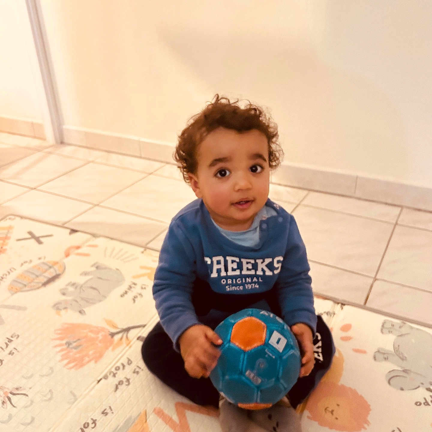 Jackson a rejoint le concours — aidez-le/la à gagner de superbes lots ! ball, blue_sweater, casual_clothing, child, curious, curly_hair, cute, floor_mat, home, indoor, looking_up, person, play, play_mat, sitting, smiling, tile_floor, toddler, toy, young_child
