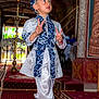 child, boy, traditional_clothing, blue, white, hat, peace_sign, carpet, indoors, decorative, pattern, portrait, smiling, person, fashion, cultural, shoes, standing, expression, celebration