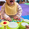 Alana is registered to the contest to win money with this photo: baby, child, infant, smiling, happy, portrait, indoor, playroom, toy, musical_toy, drum, keyboard, bib, headband, curly_hair, sitting, hands, colorful, play_mat, face