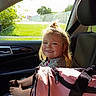 child, car_interior, seat, smile, hair, sunlight, window, plastic_bag, pink_bag, clothing, seatbelt, portrait, happy, person, daylight, hair_tie, hand, car_door, greenery, outside_view