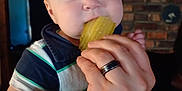 Lewis is registered to the contest to win money with this photo: adult_hand, baby, blue_eyes, brick_wall, child, closeup, cozy, cute, expression, feeding, fingers, food, hand, indoor, infant, person, pickle, portrait, ring, striped_shirt