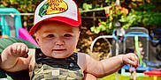 Lewis joined the competition — help win amazing prizes! baby, child, hat, cap, camouflage, overalls, outdoor, sunlight, smile, fist, person, playground, greenery, blurred_background, summer, daylight, cute, holding, happy, infant