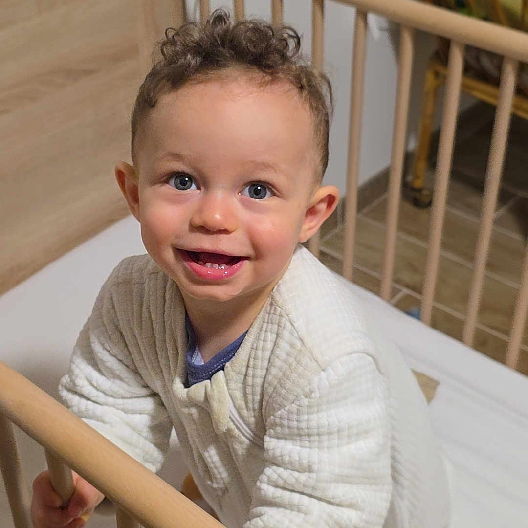 Louméo a rejoint le concours — aidez-le/la à gagner de superbes lots ! baby_clothes, bed, blue_eyes, child, crib, curly_hair, cute, floor_tiles, furniture, grip, happy, home, indoor, infant, nursery, person, quilted_outfit, smiling, toddler, wooden_crib