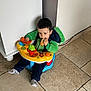 baby_seat, blue_seat, bread, candid, casual, child, cute, dinosaur_hoodie, floor_tiles, green_hoodie, home, indoor, person, playing, sitting, snack, toddler, toy, toy_phone, young_child
