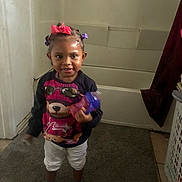 Ahlani joined the competition — help win amazing prizes! toddler, child, indoor, bathroom, carpet, laundry_basket, sippy_cup, hair_bows, socks, sweater, white_pants, smiling, person, portrait, standing, toy, curtain, floor, wall, door