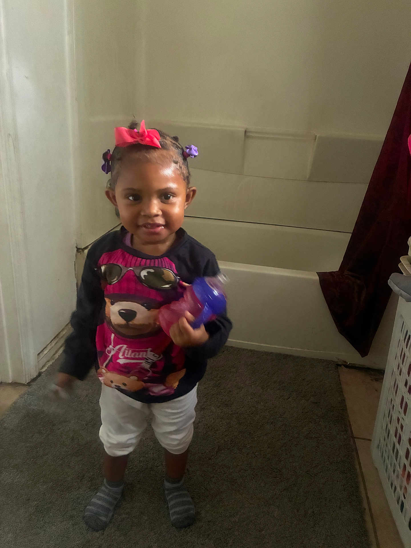 Ahlani joined the competition — help win amazing prizes! toddler, child, indoor, bathroom, carpet, laundry_basket, sippy_cup, hair_bows, socks, sweater, white_pants, smiling, person, portrait, standing, toy, curtain, floor, wall, door