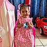 child, girl, pink_dress, smiling, indoor, toy_car, play_tent, chair, curtains, hair_buns, bows, tile_floor, white_shoes, packet, happy, cute, portrait, young_child, home, playtime