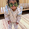 toddler, child, curly_hair, top_bun, floral_outfit, long_sleeves, pants, socks, indoor, wooden_crib, floor_mat, playful_expression, green_glasses, novelty_glasses, person, young_child, standing, home_interior, cute, smiling