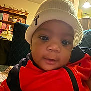 Treyvion is registered to the contest to win money with this photo: baby, child, hat, red_jacket, face, indoor, furniture, shelf, lighting, portrait, cute, knit_hat, warm_clothing, young_child, curious, cozy, home, person, closeup, expression