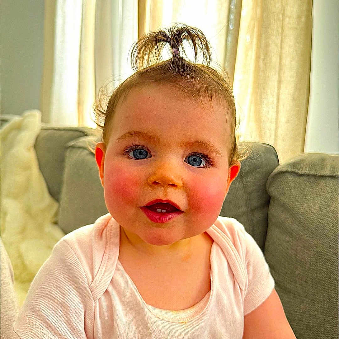 Juliana participe au concours pour gagner de l'argent avec cette photo : baby, blue_eyes, child, clothing, couch, curtains, cute, face, happy, home, indoors, infant, person, ponytail, portrait, rosy_cheeks, sitting, smiling, soft_lighting, white_shirt