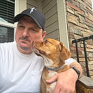 Maple joined the competition — help win amazing prizes! man, dog, portrait, selfie, outdoor, porch, pet, companion, kiss, affection, brown_dog, white_shirt, baseball_cap, beard, collar, stone_wall, window, railing, wicker_furniture, daylight