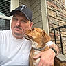 man, dog, portrait, selfie, outdoor, porch, pet, companion, kiss, affection, brown_dog, white_shirt, baseball_cap, beard, collar, stone_wall, window, railing, wicker_furniture, daylight