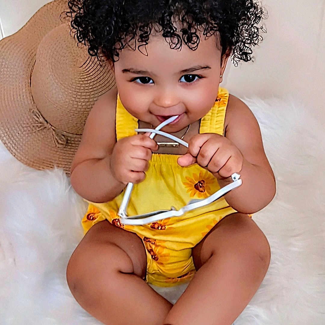 Monroe'Blu is registered to the contest to win money with this photo: accessory, baby, barefoot, brown_sandals, child, curly_hair, cute, fashion, fluffy_surface, happy, hat, indoors, person, playful, portrait, sitting, smile, sunglasses, toddler, yellow_outfit