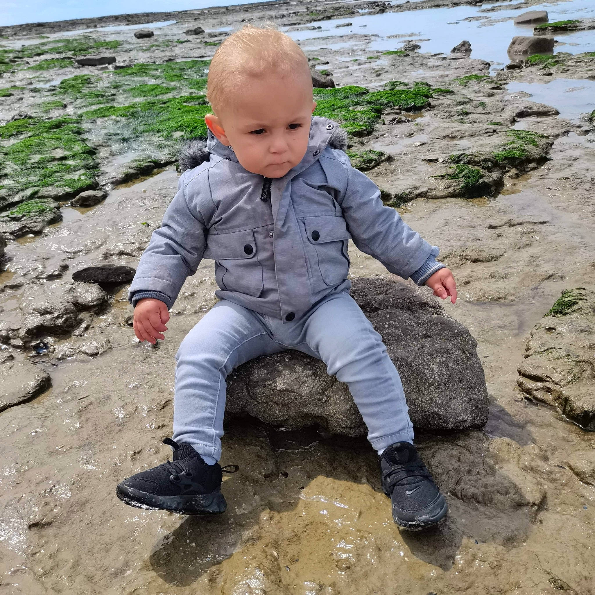Sohan a rejoint le concours — aidez-le/la à gagner de superbes lots ! baby, baby_toddler_clothing, bedrock, coast, fun, grass, head, landscape, mud, people_in_nature, person, plant, play, recreation, rock, sand, sitting, sneakers, soil, toddler