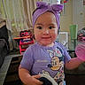 baby, child, infant, smiling, portrait, indoors, purple_headband, bow, purple_shirt, cartoon_shirt, toy, spiderman, pink_pom_pom, living_room, table, chair, curtains, door, mirror, home_decor