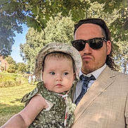 Nova is registered to the contest to win money with this photo: baby, adult, man, child, outdoor, tree, sunglasses, hat, dress, green, nature, sunlight, grass, park, person, holding, portrait, daytime, leafy, casual