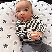 Avishay is registered to the contest to win money with this photo: baby, infant, striped_clothing, gray_pants, star_pattern, cushion, face, hands, curious, wide_eyes, indoor, cute, child, portrait, sitting, relaxed, soft_fabric, cozy, young_child, casual_wear