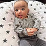 baby, infant, striped_clothing, gray_pants, star_pattern, cushion, face, hands, curious, wide_eyes, indoor, cute, child, portrait, sitting, relaxed, soft_fabric, cozy, young_child, casual_wear