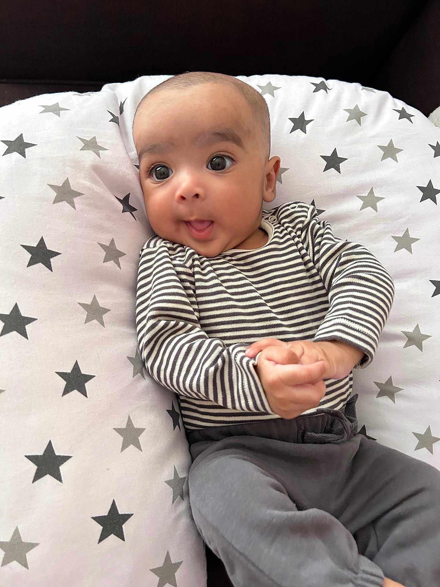 Avishay is registered to the contest to win money with this photo: baby, infant, striped_clothing, gray_pants, star_pattern, cushion, face, hands, curious, wide_eyes, indoor, cute, child, portrait, sitting, relaxed, soft_fabric, cozy, young_child, casual_wear