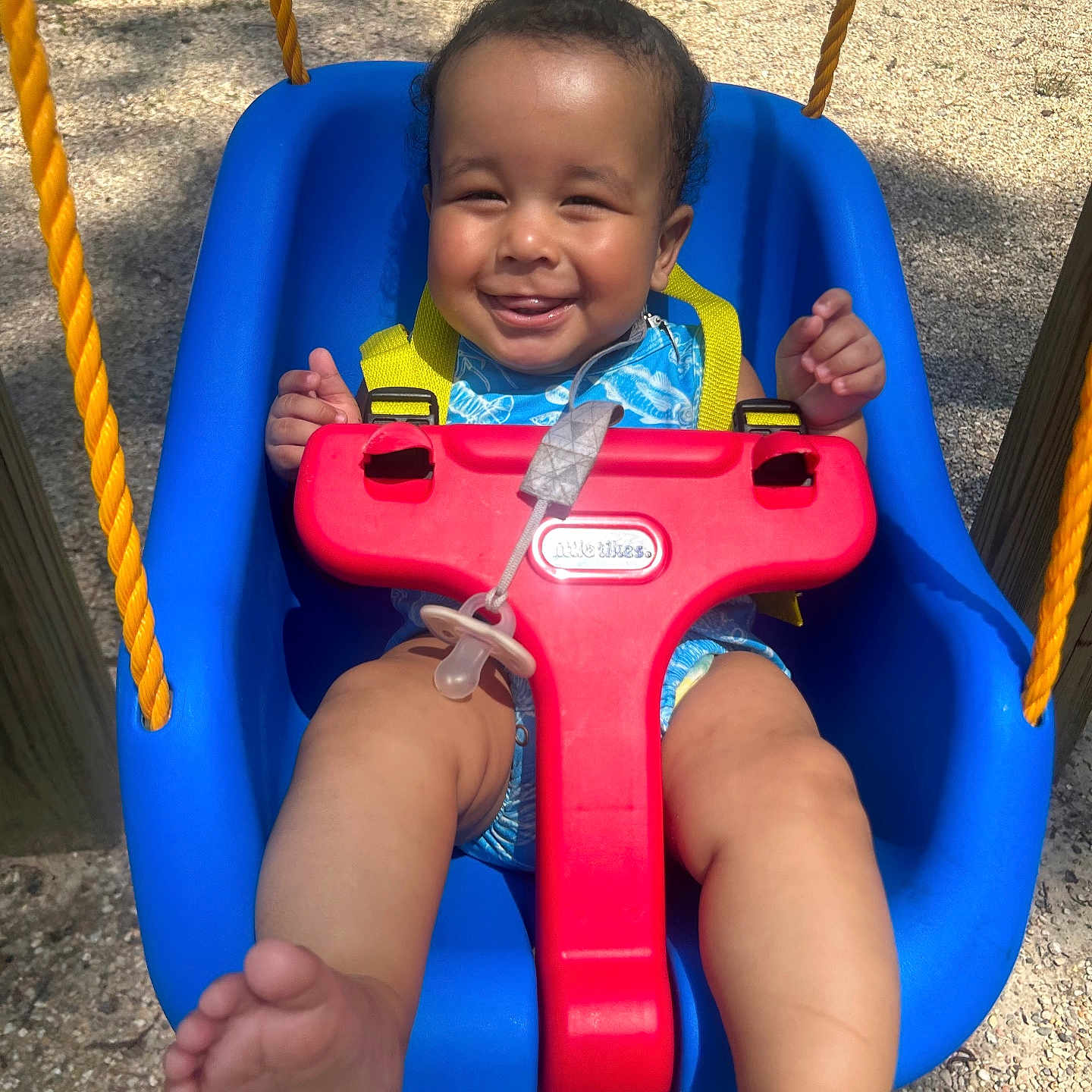 Zain joined the competition — help win amazing prizes! baby, child, swing, playground, smile, happy, outdoor, sunlight, blue_swing, yellow_straps, infant, legs, feet, red_safety_bar, pacifier, tree, gravel, play_equipment, cute, fun